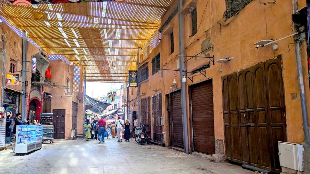 a dark and somewhat unnerving Marrakech market street. Although it's quite wide, and you can see the exit. There's no one close to you, and the shop doors are all closed on the right side. There are a few shops open on the left. There's a removable rooft, made out of bamboo maybe? But the sun can still peak through. After the opening, it's a lot brighter, and you can see people walking there. 