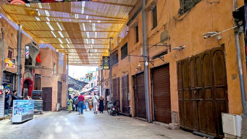 a dark and somewhat unnerving Marrakech market street. Although it's quite wide, and you can see the exit. There's no one close to you, and the shop doors are all closed on the right side. There are a few shops open on the left. There's a removable rooft, made out of bamboo maybe? But the sun can still peak through. After the opening, it's a lot brighter, and you can see people walking there. 