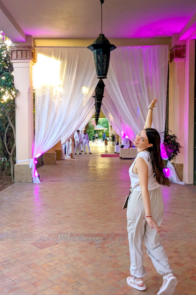  a woman walking down a corridor with her hands wide open, one is up in the air, the other is down by her legs as she tilts her head back to the camera as though showing off what she's walking towards - two swooping white curtains hang from the ceiling. She's wearing a white muscle tank top and loose fitting pants that are tied at the ankle by a drawcord, she's wearing white teva sandals