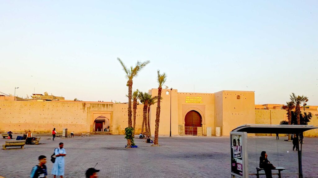 the outside walls of the marrakech medina, old city. They aren't tall, have a yellowish hue during golden hour, a large round doorway to enter through. There are three lonely palm trees in the concrete courtyard leading up to the walls.