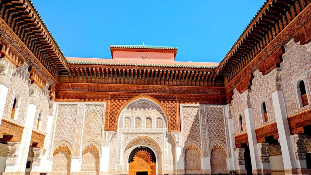 the inner courtyard on a beautiful sunny day. You see three walls of the courtyard, and the sky beaming in a bright blue above. The sun casts a shadow on the right wall but the other two remain bright as ever. They are tiles in white and soft terracota colours. There's a large wooden doorway in the middle wall, this brings you to the chapel. Archways on the side walls lead you to the inner rooms