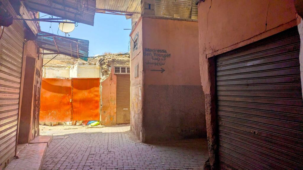 a creepy street in the market of marrakech. All the doors are closed (they look like brown painted garage doors), on  one of the walls, you can see it painted Medressa Ben Youssef with an arrow pointing to the right, so you know you're actually supposed to be here. It's a tad less creepy because the ripped fabric roof ends, and you can clearly see the blue sky and sunlight coming in