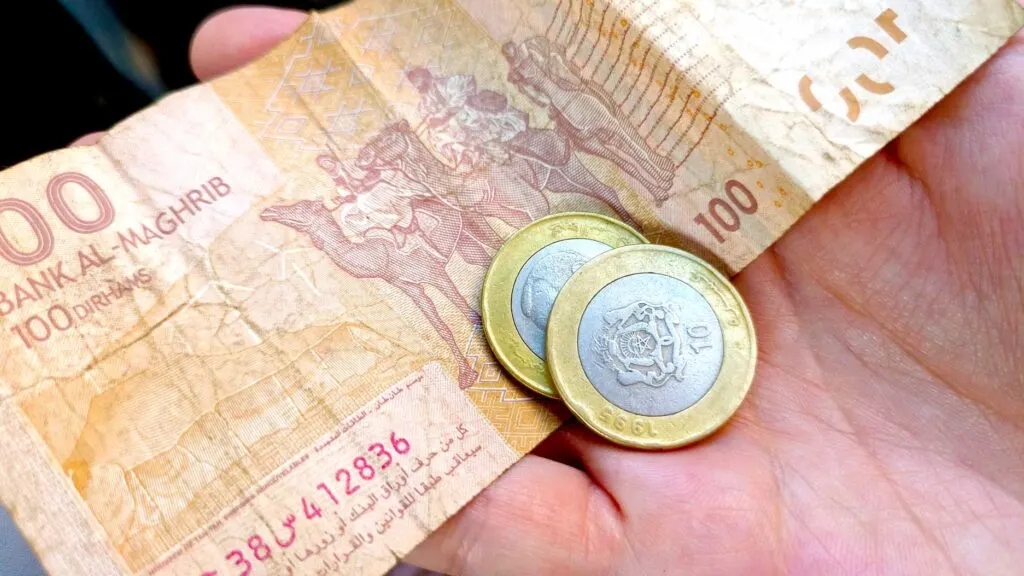 an example of Moroccan currency: dirham. A woman is holding a 100 dh bill and two 10 cent coins in her hand. The bill is a rusty orange colour with camels on it. The coins are silver on the inside with the moroccan emblem and a gold rim