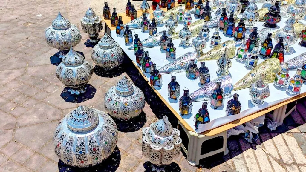 a display of moroccan lanterns outisde at a market in marrakech. Two rows of lanterns sit on the floor, they are the same style but vary in size.  Behind them on a raised platform sits smaller lantern options, these are different with glass and different lantern designs