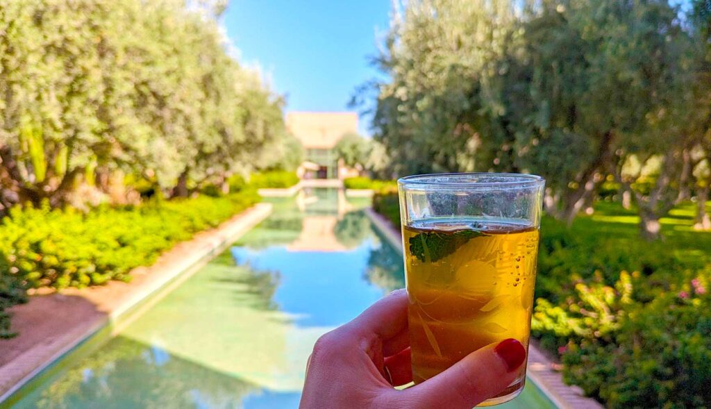 a woman's hand is holding a small glass of moroccan mint tea, you can still see the fresh mint leaf floating in the tea, which is a yellow brew. Behind the tea is a blurred moroccan scene of a stream of water with gardens on either side, leading to a pavillion in the distance. The water is so still that you can see the reflections of the trees and the pavillion. Its a sunny day, casting a yellow hue on the photo.