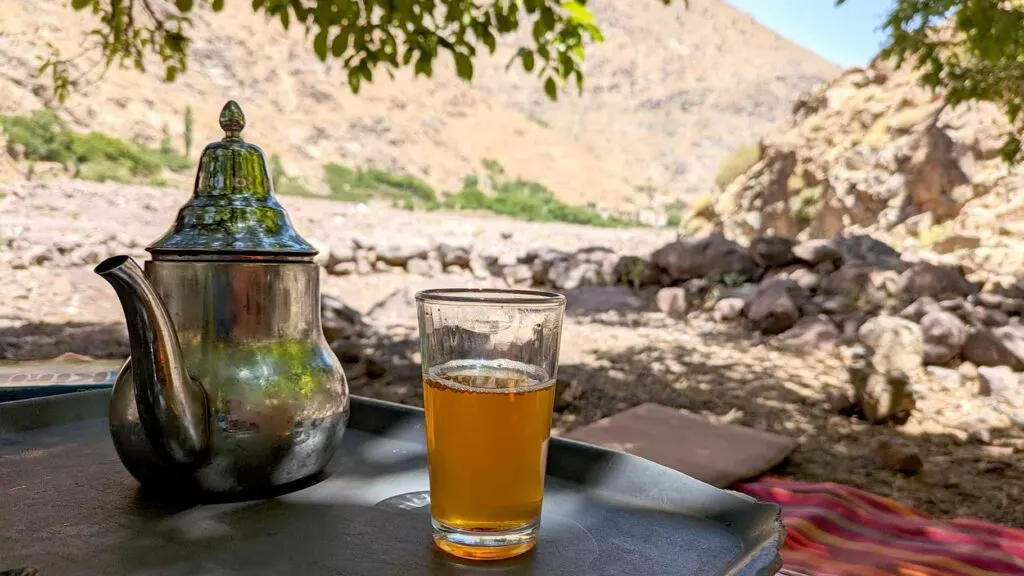 a moroccan teapot with a filled glass with tea next to it on a table. You can see a red carpet on the ground behind it. And you can see that you are in the mountains. There isn't anothe rperson in sight, theres some greenery but on the mountains its all rocks. You can only see a sliver of the sky peaking through two mountains.