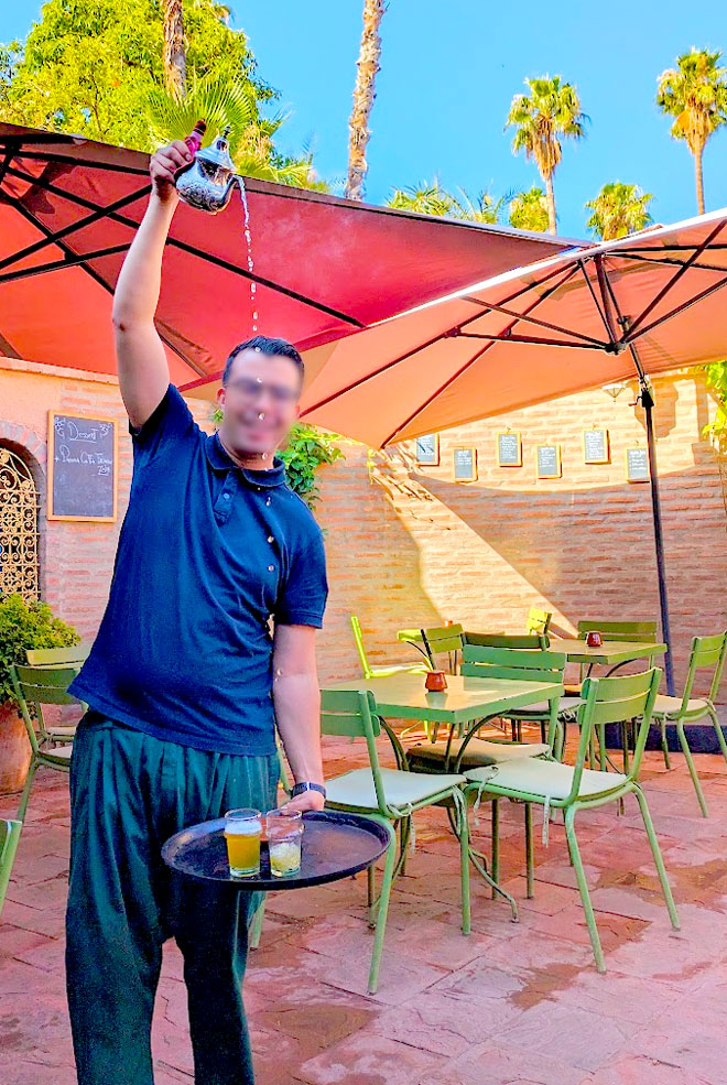 the best mint tea pour in marrakech at the marjorelles cafe. a man is holding train with two cups in one hand, extended below his waist. Then high above his head is his second hand hold the teapot pouring tea into the cups. You can count the droplets of tea as it falls into the cups, and you still see the steam leaving the hot tea