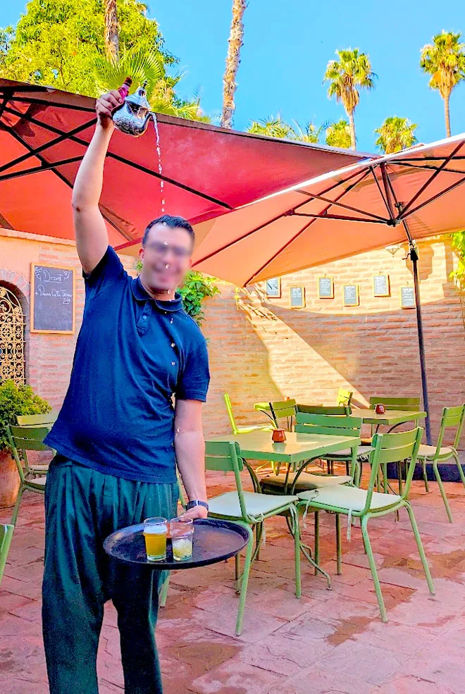 the best mint tea pour in marrakech at the marjorelles cafe. a man is holding train with two cups in one hand, extended below his waist. Then high above his head is his second hand hold the teapot pouring tea into the cups. You can count the droplets of tea as it falls into the cups, and you still see the steam leaving the hot tea