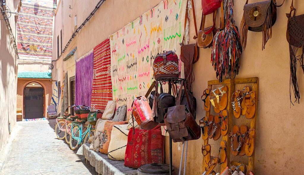 a narrow street in the middle of tall stucco pinkish coloured walls. Theres a small wooden doorway at the end. On the taller wall, there s an array of rugs hung up for display along with some purses and shoes for sale