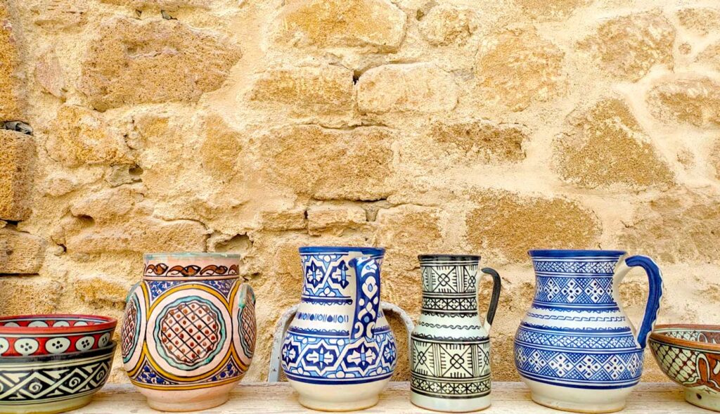 six examples of morroccan pottery on display on a table against an old stone wall. There are two bowls that are cut off on either end of the photo. The middle has four vases with moroccan patterns, two of which are blue and white classics