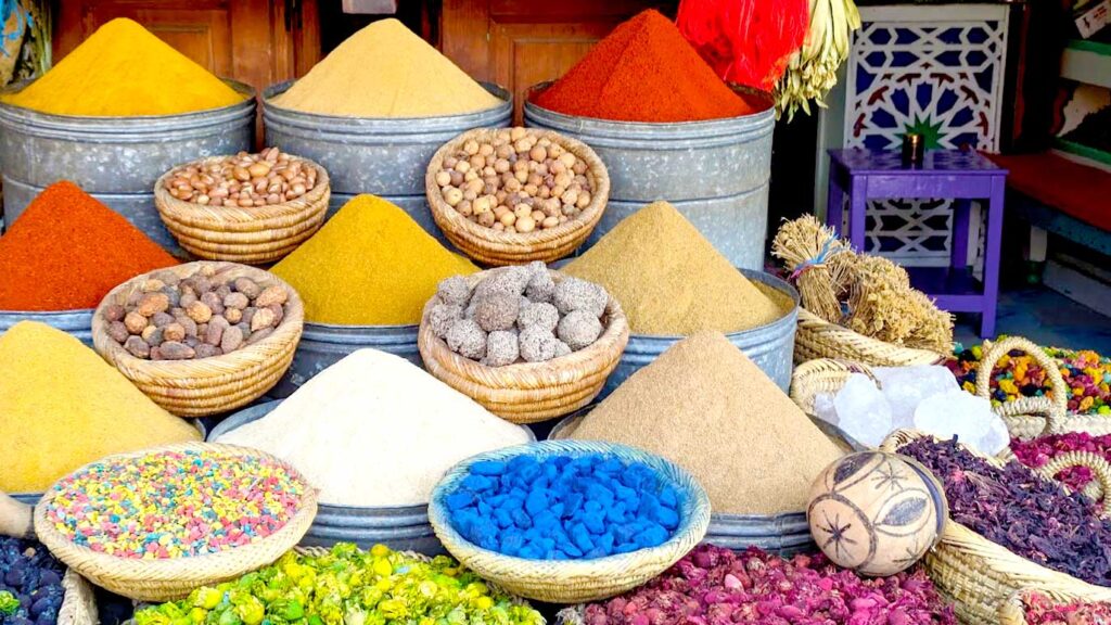 a beautiful display of moroccan spices in Marrakech. There are four rows of three vats of spices that have been piled high and shaped like a pyramid with a peak at the tip. In between each spice pyramid is a bowl of either nuts, or dried petals. The spuces are varying shades of yellow, there's a bright red spice, and there's a bright vibrant blue powder as well creating a very colourful image 