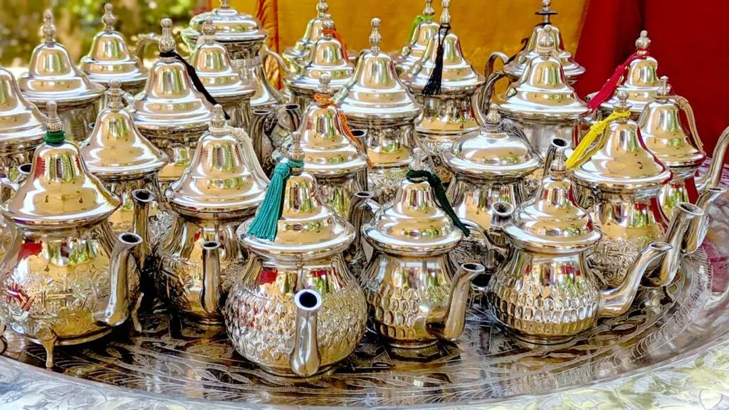 25 silver moroccan teapots for sale sitting on a silver tray table. Most of an imprinted design, some rest on silver legs, and others have a tassel at the top of the teapot cover. They all vary in sizes