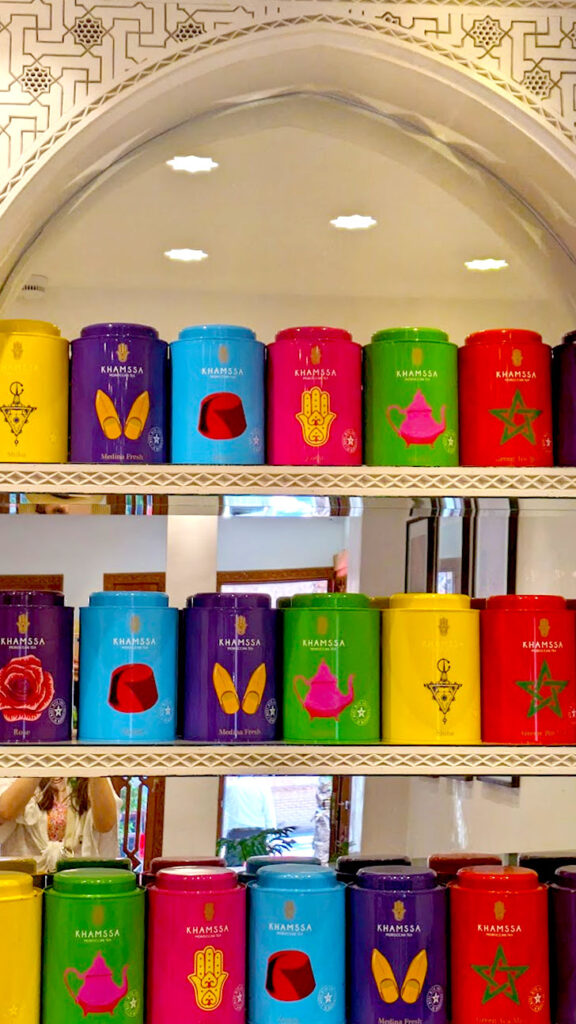 three shelves sit in front of a mirror, that is rounded at the top, with a patterned designed on the wall above it. On the three shelves are tins of Khamssa tea. There are seven different types of tea, each with a different colour tin and symbol on the front. Some of the symbols are babouches, a fez hat, a moroccan teapot, a Moroccan star and a hamsa 