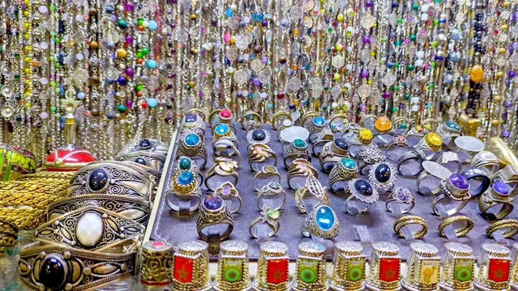 moroccan jewellery on display at a souvenir shop. On a display case is a tray with silver rings, some stones and gems. Next to it sits 4 rows of bracelet bangles with a large stone.  Behind the display case is the wall filled with more silver necklaces dangling.