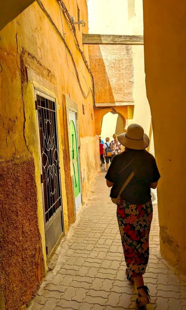 a vertical image: A woman is trailing behind a group of people, as they walk through a street in the Marrakech medina. Although it's bright, there isn't a roof letting in the warm yellow sun, the walls are VERY tall. And besides for the group, there's no one else here.