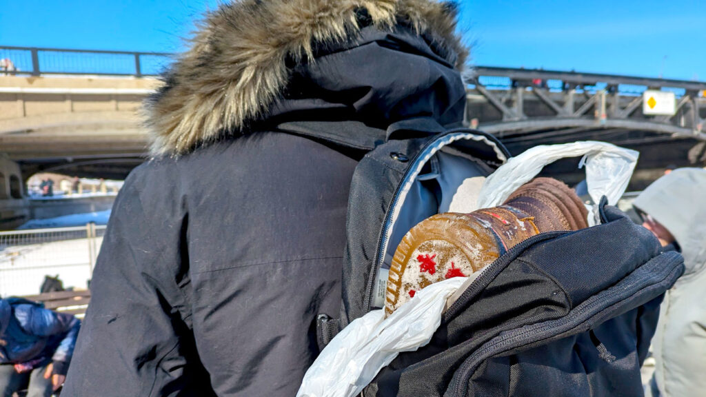 a man in a winter jacket photographed from behind. He takes up most of the photo, with the open backpack on his back. Inside the backpack you can see a white plastic bag lining the bag, and inside, a the bottom of a pair of winter boots. 