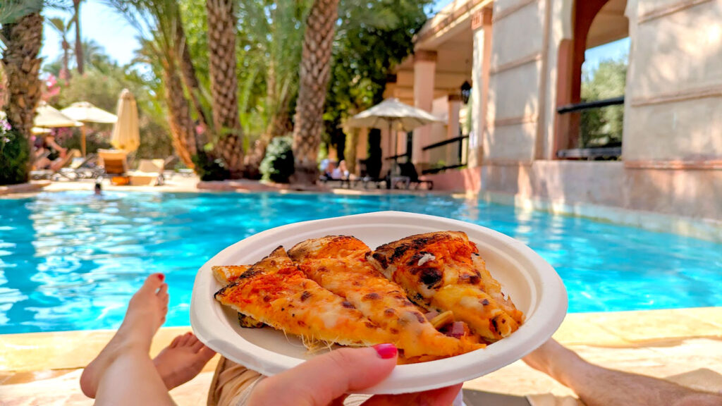 a hand holding up a paper plate filled with slices of cheese pizza. But she's not at a restaurant, you see her bare red painted toes, with a blue pool and palm trees behind her. 