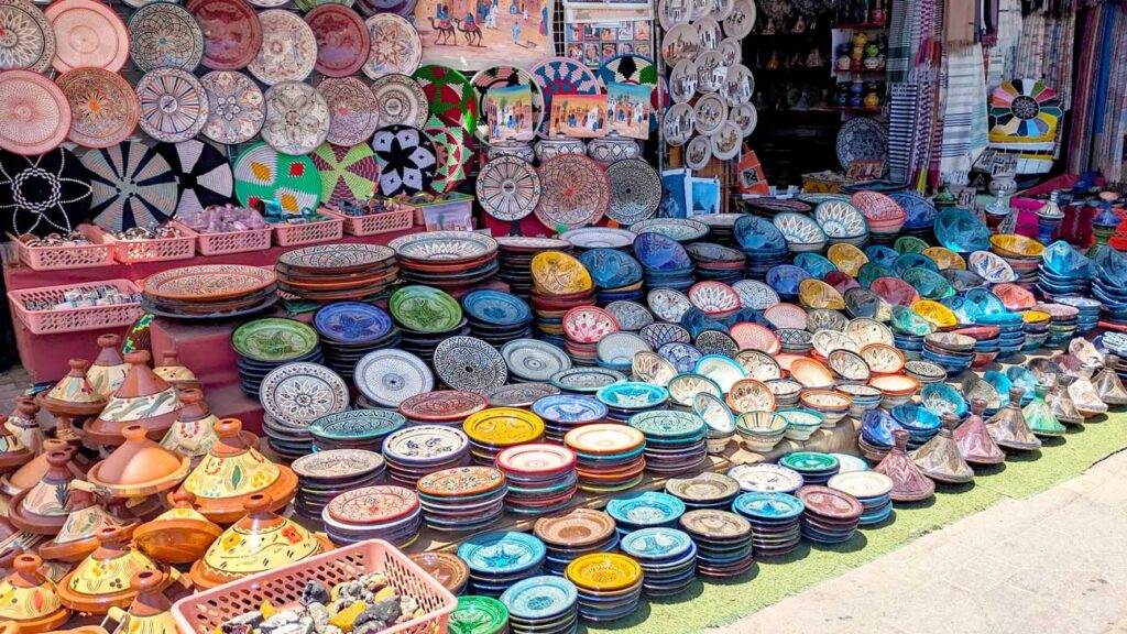 an outdoor vendor display of ceramic plates and tagines in the markets of marrakech. Every plate is a unique pattern and colour, there are probably over 100 plates here. Smaller plates are stacked in rows on the ground, and there are larger ones on a table behind, and even more hung up on display on the wall