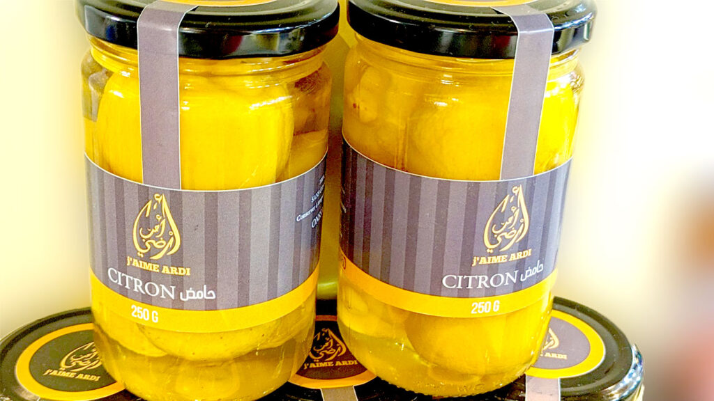 Two jars of preserved lemons are sitting on top of three more jars of preserved lemon at a store, the background is blurred