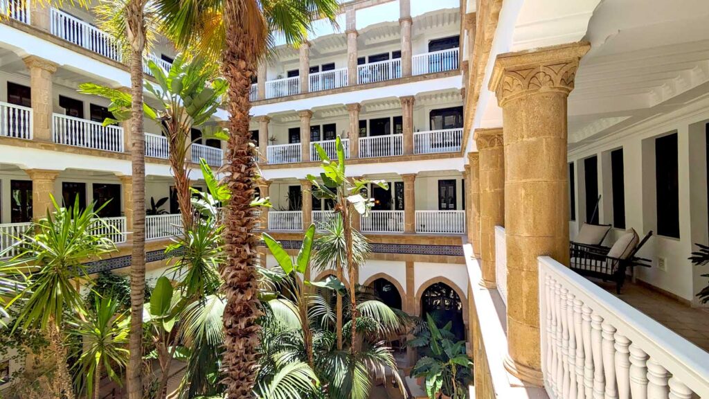 an example of a riad hotel in Essaouira Morocco. Looking at the inner couryard form the second floor of a riad. There is a railing leading your eye to the right, there are doors a bit further inside the outdoor corridor. These outdoor corridors, with railings, go all the way around an inner courtyard, and up several stories - although you can't see how many. In the middle is a beautiful garden, you can only see the tops of some shorter palm trees, and the trunk of another one that the top is halfway cut off it's so tall. 