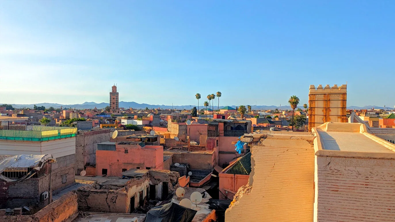 the incredible rooftop view at sunset from shtatto rooftop. Right below are some ruined buildings, with trash bags, satellite dishes, and crumbled walls, but beyon that are a few palm trees that tower above the rooftops, at the same height as one mosque in the distance. There's a warm yellow glow to all the buildings. But far away, you see the outline of some mountains, those are still a nice shade of blue. There isn't a cloud in the sky