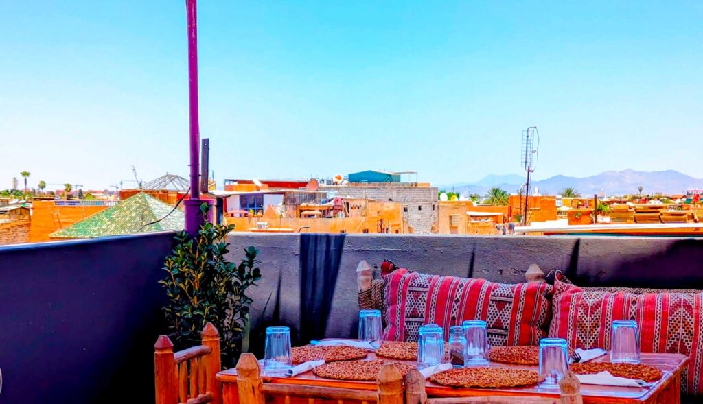 a top marrakech things to do: eat lunch on a rooftop in the medina. There's a table for four set up against a rooftop barrier. There are moroccan printed pillows for comfort. Behind the barrier, you can see other rooftops of marrakech and finally, the mountains in the distance.