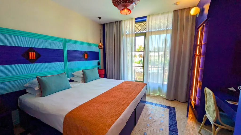  a very moroccan inspired room in the family oasis at the club med marrakech. The head board is bright blue and dark blue stripes. A king bed is in the middle with white bedding, and an orange accent throw on the end. There's a large balcony doors, covered by sheer curtains. The floor is all tiled. 