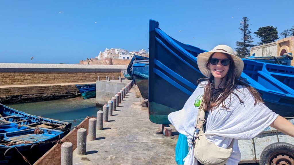 a woman showing off her uniqlo round min shoulder bag. Attached to the strap is a mini bottle of purrell. She's wearing a white wrap, and a straw hat. She's standing in front of a vibrant blue boat and off in the distance is the walled white city of essaouira 