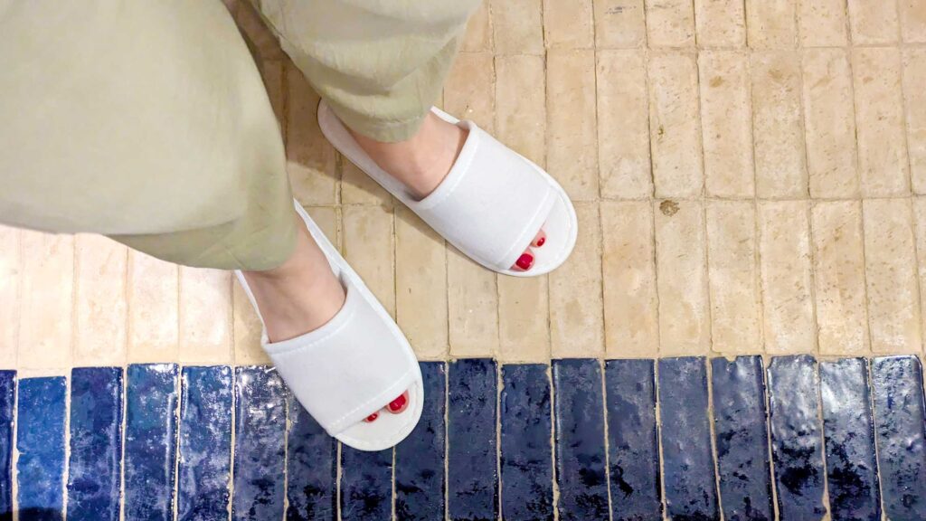 a top down view of a womans feet standing on tiles. The bottom are navy glazed tiles, while she's mostly standing on unglazed rougher beige tiles. She's wearing white hotel slippers, you can see her red nail polish peak through the top. She's wearing beige cargo pants