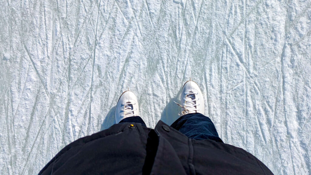 a top down view of a woman's figure skates on ice. The ice has a bunch of lines crossing it, clearly been skated on before. She's wearing navy corduroys, and you can see the bottom of her thick black winter jacket. 