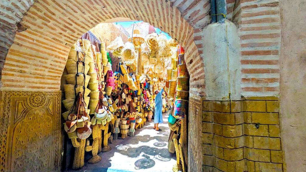 a doorway where you can see straw bags and hats lining the wall, a man is trying to reach one of the bags that are higher up. The sun light is shining in beautifully creating a surreal ambiance to the picture, illuminating the straw bags as the perfect souvenir from morocco