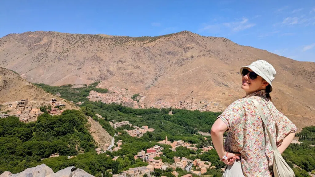 a woman stands triumphantly at high above a valley on a mountain side as other mountains soar in the sky behind the valley. She's wearing a sun hat, with sunglasses.