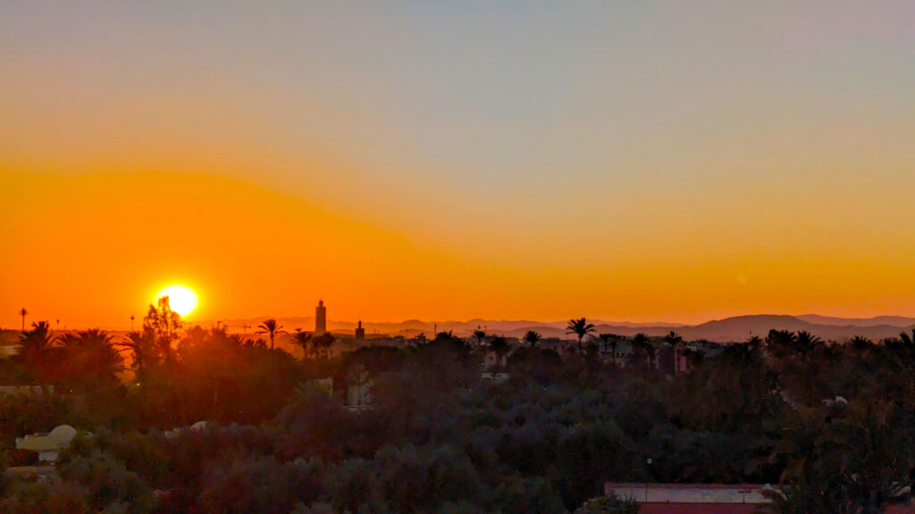 the view of sunset from the club med over marrakech. The sun is almost behind the horizon, creating a rich orange colour before it turns into a grey blye higher up. It also casts a a shadow over marrakech, you can see trees, but the buildings mostly blur into each other, except for a mosque tower that rises above the skyline And different shade of shadows create the mountains along the horizon