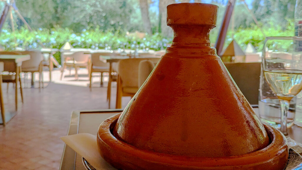 a clay unpainted tagine sits closed on a table, no one knows yet what goods are inside! Behind the tagine is an empty tables at the El Kebir restaurant. It's open with a cover for shade. 