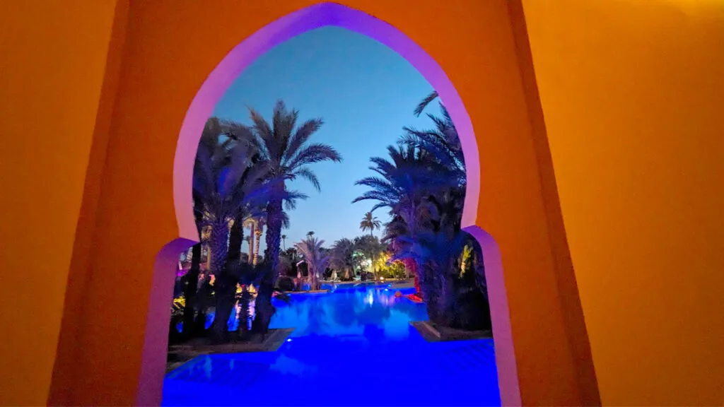 a semi circle rounded archway in morocco, peaking out onto the pool oasis at the club med morocco at night. There are shadowed palm trees on either side of the vibrant royal blue lit pool