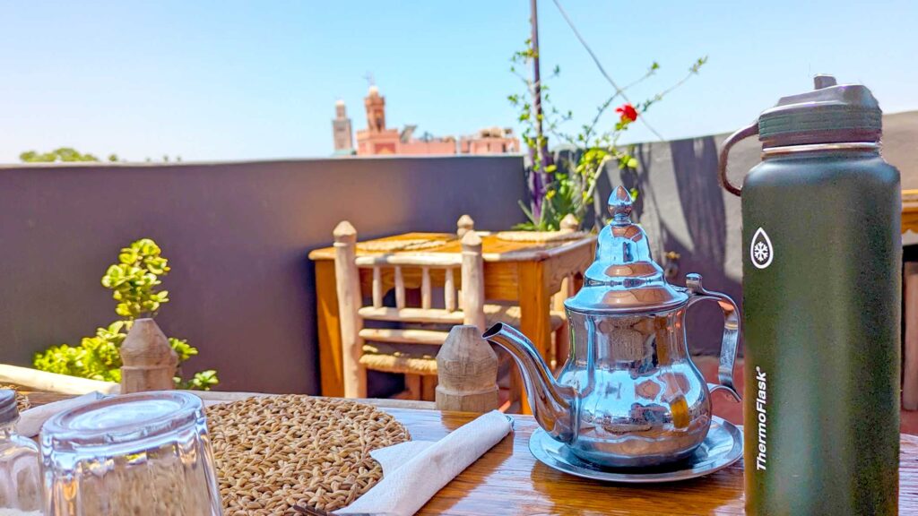 a rooftop restaurant in marrakech. A table is cut off, but you can see one place setting, with a moroccan tea pot next to the forks, and a thermoflask water bottle on the edge of the table. Above the rooftop ledge, you can see two mosques. 