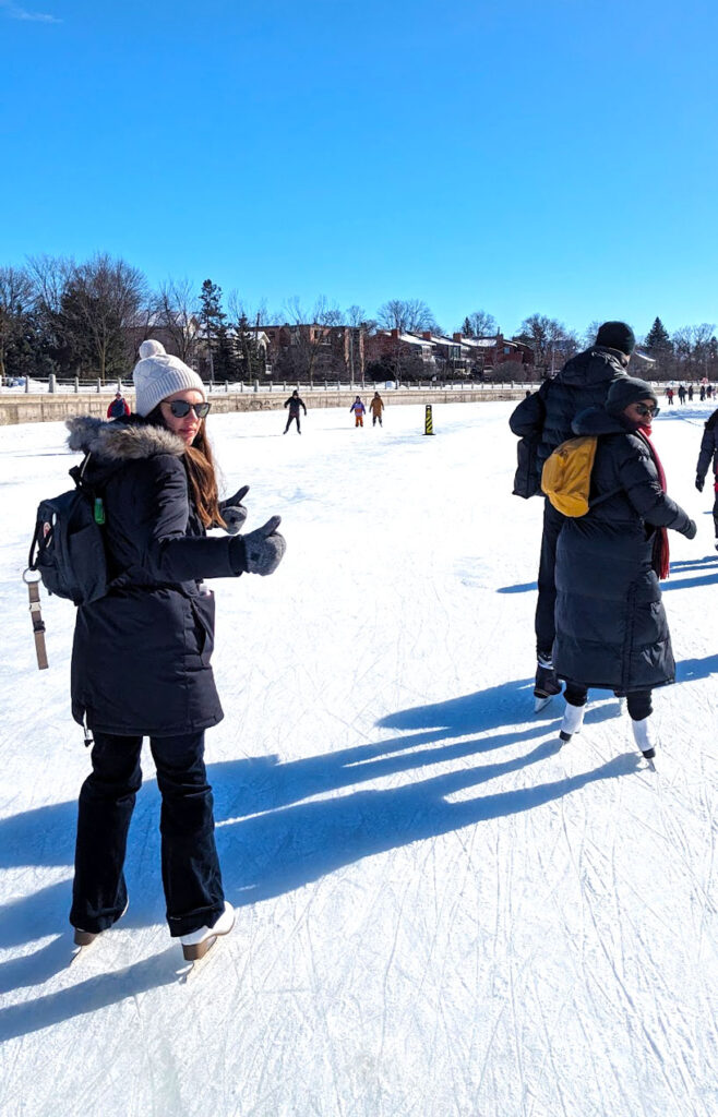 a woman stands on ice, wearing skates and full winter gear, she's looking back at the camera with a serious look on her face but giving two thumbs up. It's a very sunny day, the ice is white with a bit of snow covering, and there isn't a cloud in the sky.