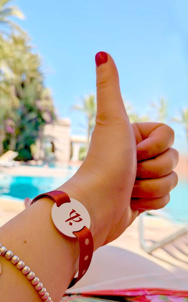 a woman's arm is held up in the middle of the frame with a thumbs up, she has red nail polish on. On her arms are two bracelets. The first one is a burnt orange ribbon band holding a plastic white circle in the middle with the club med logo on it. And then further down her arm is a silver beaded bracelet. 

It's a bright and sunny day, you can make out a bright blue pool behind her hand