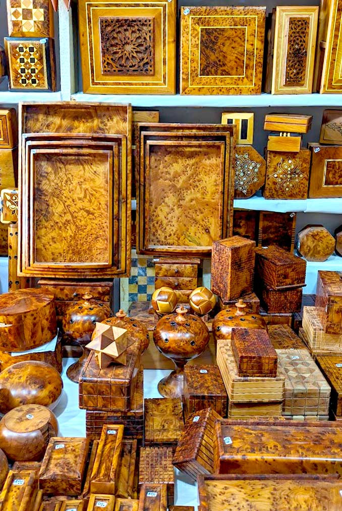 the entire frame is filled with different hand crafted items made from thuya wood in essaouira. On a table sits four ros of boxes, and circular containers - there's no organization. there are two sets of nesting trays leaning on the wall shelf behind it. On the shelfs are even more boxes.  