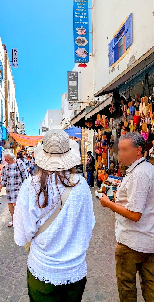 a tour guide turns around to speak to the woman next to him, her back is completely to the camera, she's wearing a shawl and a straw hat as they walk through the lively market in essaouria with blue andwhite buildings and souvenirs on display outside