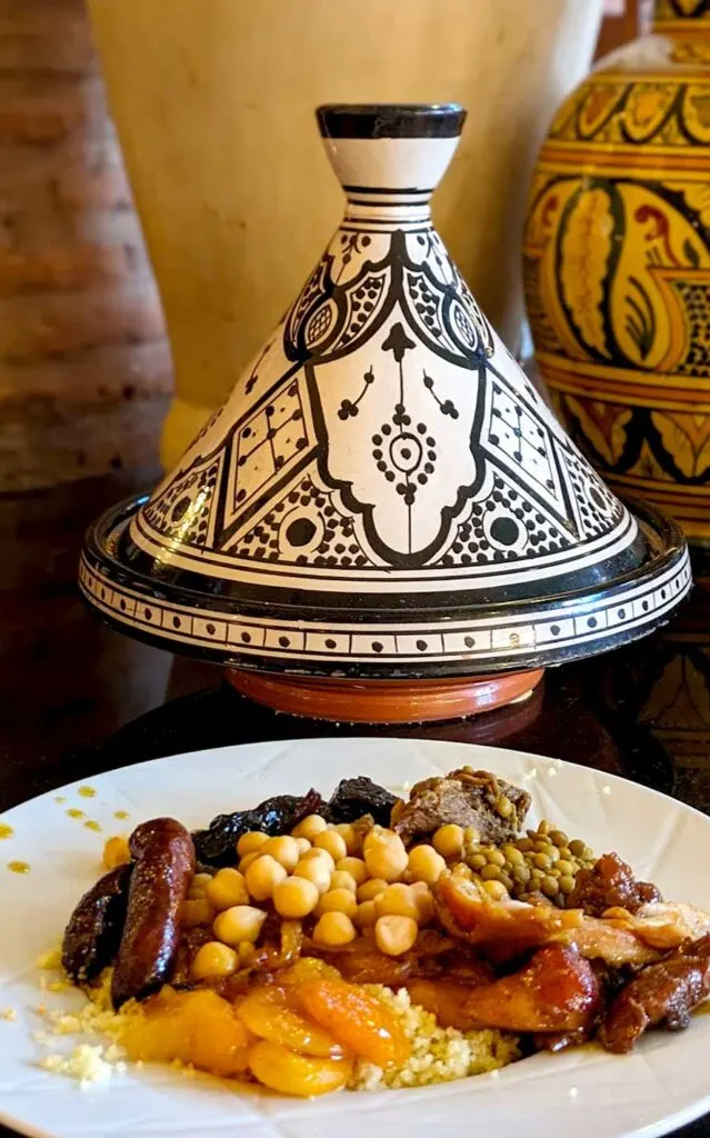 a completely filled place of Moroccan cuisine. You can see cooked dried apricots, couscous, a beef merguez, chickpeas, chicken and lentils. Behind the plate is a beautifully crafted decor tagine. It's a black and white geometric Moroccan pattern