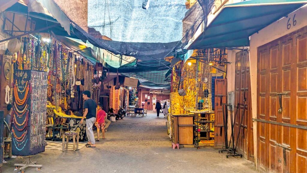 A less busy street in the Marrakech medina. There's a thin black cloth roof, that you can still see the sky through. The right side of the street, are all closed wooden doors. On the left, there are some vendors selling jewellery. In the distance you can see some tourists walking