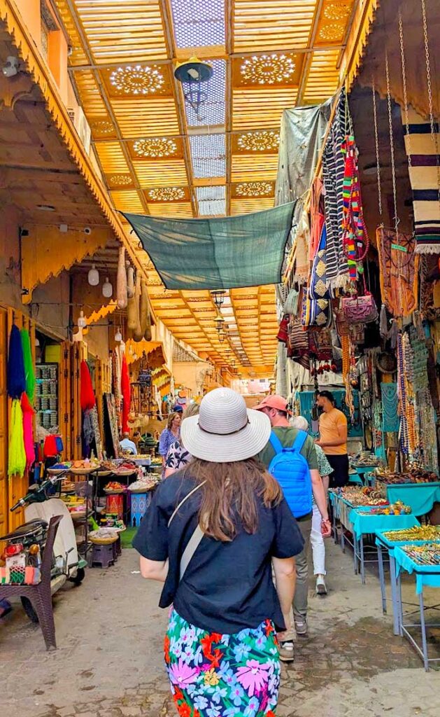 a woman's back is in the middle of the frame as she walks through the market street in marrakech. She's wearing a straw sun hat, a black tshirt, and a floral skirt. There is a roof on top of the market, that still lets in some natural sunlight. On either side of the alley are shops with vendors trying to sell their goods.