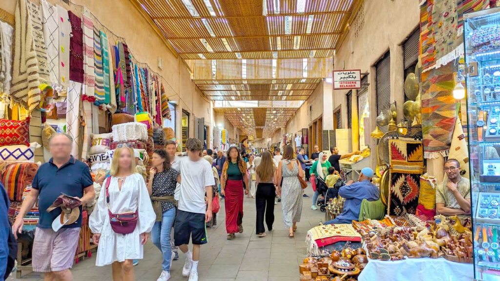 an example of one of the main bustling market streets in the Marrakech market. People are walking through a wide covered street with vendors on either side. The roof isn't completely covered, so some light is getting through the wood strips. 