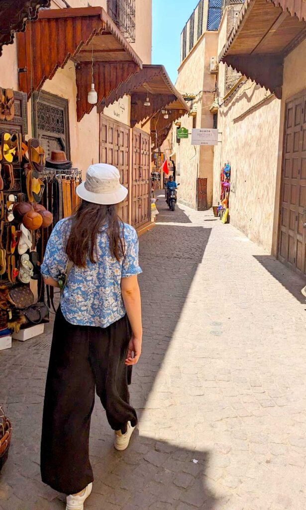 The back of a women with long brown hair walking through the quiet streets in the Medina of Marrakech. There's one shop open, the walls are tall and orange pink colour. She's wearing flowy black pants, and a loose fitting blue and white blouse as well as a cream coloured bucket hat