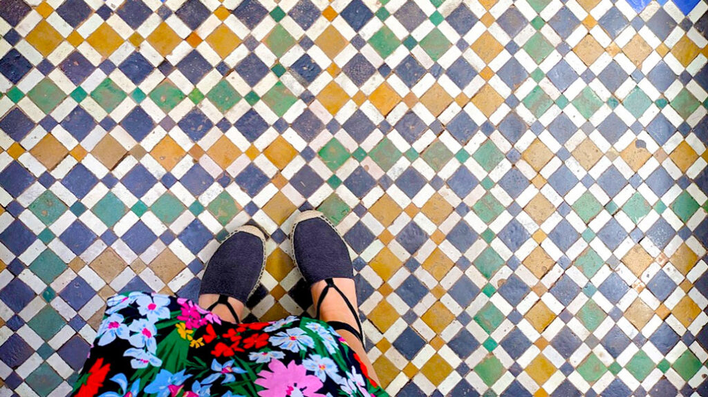 photo taken of a woman's feet from above. She's wearing black espadrille sandals, that have a rope tied around her ankle. You can't see her legs, they're covered by a long floral skirt. She's standing on zellige, moroccan tiles. It's a patterns of different shaped and coloured square tiles that are titled to look like a diamond shape. The colours of the tiles are green, navy, mustard yellow, white. 