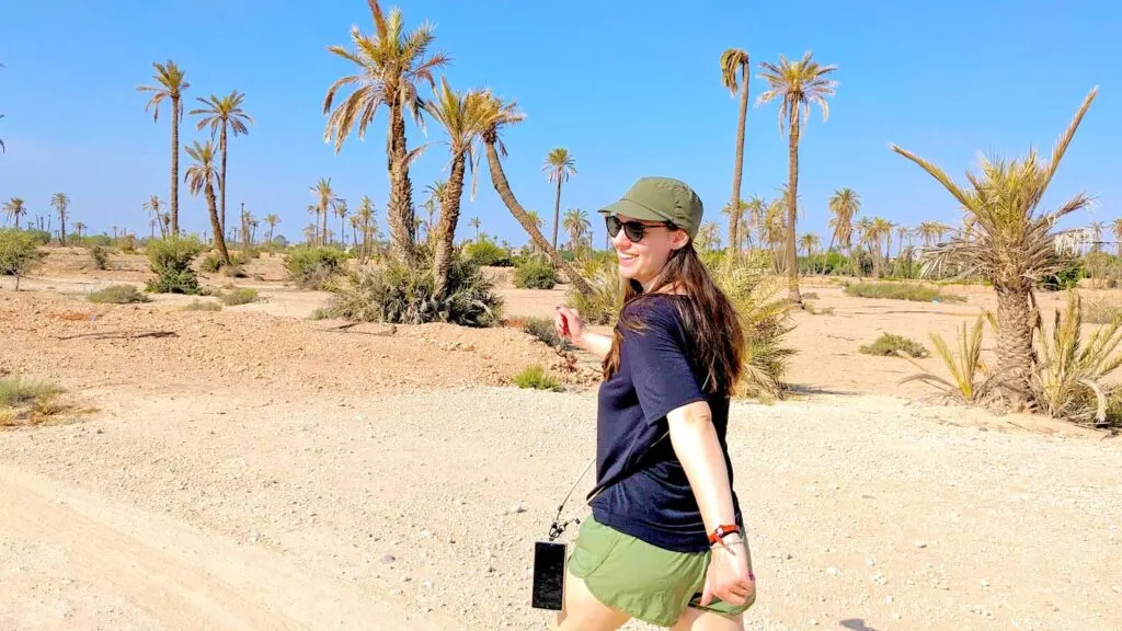 a woman briskly walking in what looks like a desert path, with a few scattered palm trees around. Her arms look like they're paused in motion, one is swung back while the other is out in front of her. She's smiling, wearing sunglasses, a thing army green baseball cap, a black workout t shirt, and army green running shorts. Her phone is also attached around her body on a rope, swinging out in front of her as she walks.