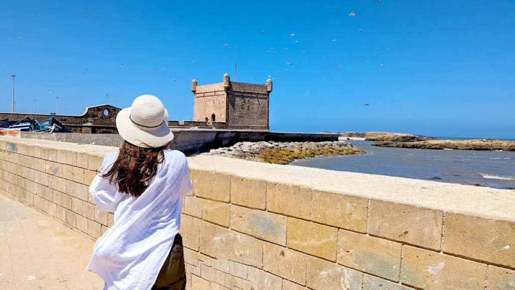 an example of what to wear in morocco as a woman. A woman stands with her back to the camera. She's in front of an old stone wall at the port, it separates the streets from the water. In the middle of the frame, behind the water leading to the short stands a reddish stone tower. 

The woman is wearing a wide brim straw hat, a white long loose kimono. You can tell its loose because it's blowing in the wind along with her hair.