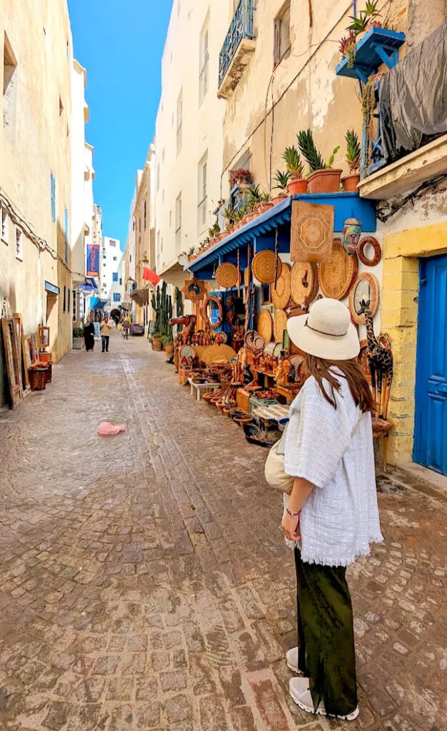 wearing pants as a woman in Morocco. A woman is standing in a street with shops on either side in essaouira, no one else is close. Her back is to the camera. You can see her long brown hair below her sun hat on her back. She's wearing a white long shawl, and long green loose fitting pants with running shoes