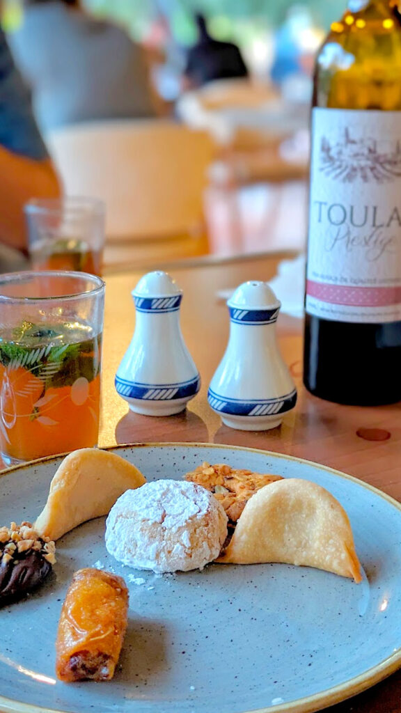 a vertical photo of a table at the el kebir restaurant. There's a plate filled with different delicious moroccan desserts, with moroccan mint tea behind it, a salt and pepper shaker and toulal moroccan wine